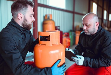 men in black jackets handing each other an orange propane tank in a warehouse